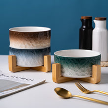 Two ceramic bowls with gradient designs on wooden stands, with a blue background and gold cutlery.